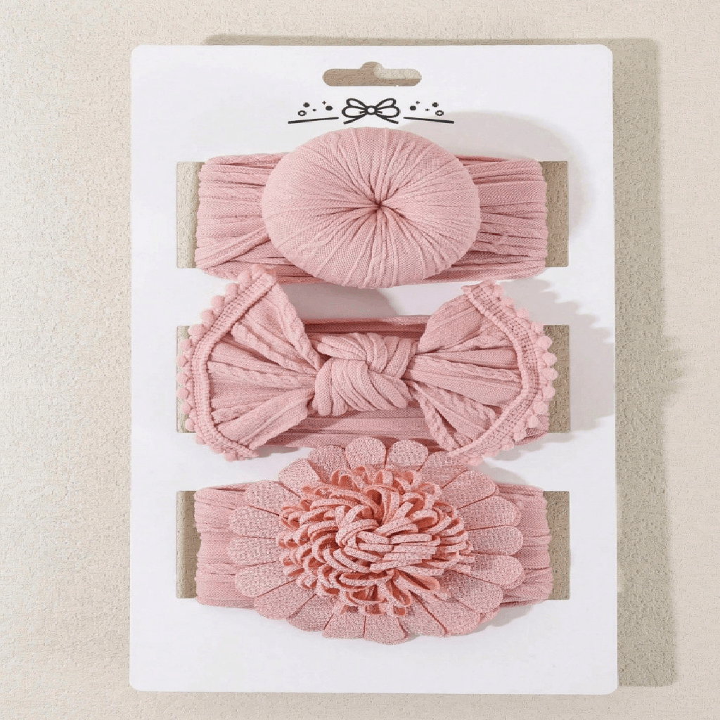 Three pink fabric headbands on a white display card, featuring a round top band, a middle band with a knotted bow, and a bottom band with a large ruffled flower, photographed on a light beige textured background.