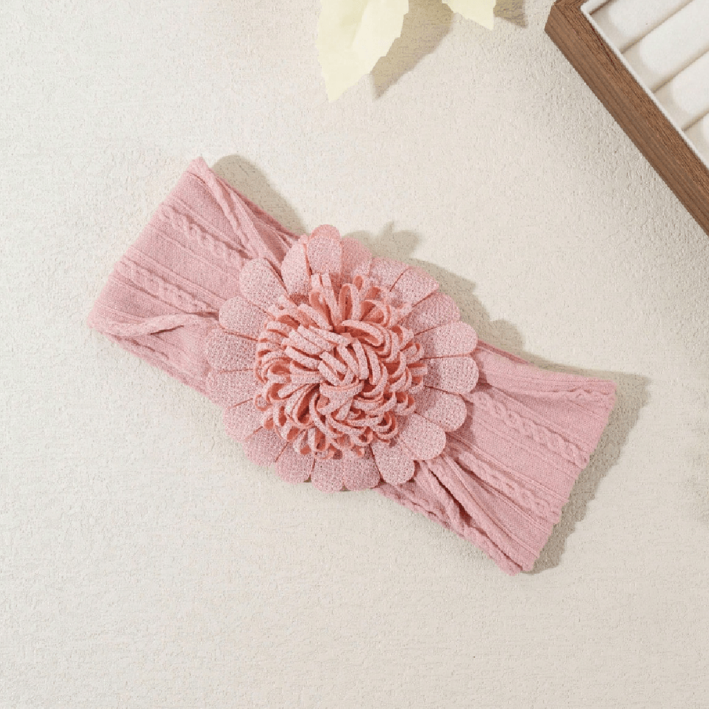A pink ribbed knit headband with a large layered fabric flower applique in the center, laid flat on a light textured surface.