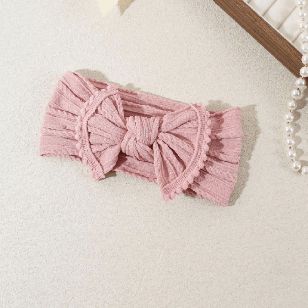 A pink ribbed knit headband with a large central bow and ruffled edges, laid flat on a light textured surface with a pearl strand and wooden frame partially visible in the corner.