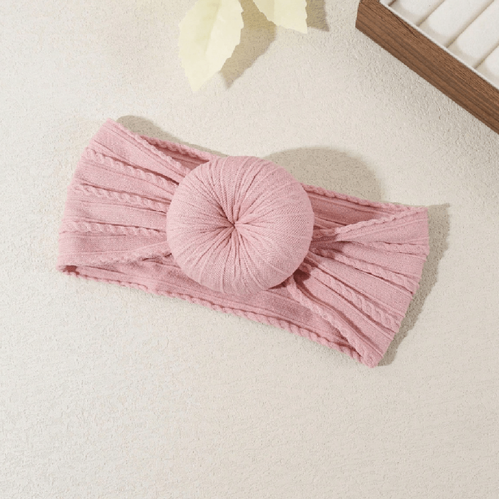 A pink ribbed knit headband with a large round knotted bow in the center, laid flat on a light cream textured surface with soft decorative props in the background.
