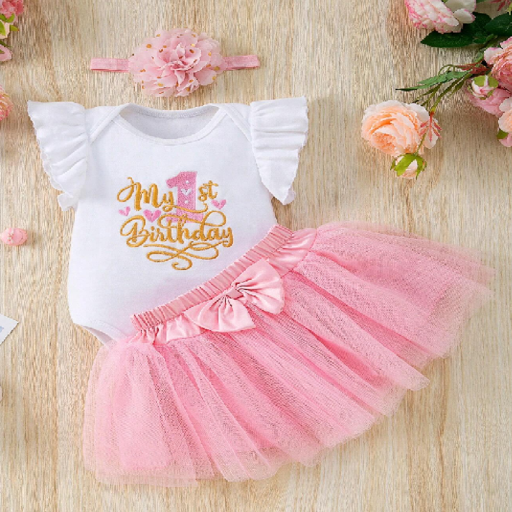 Baby outfit with 'My 1st Birthday' shirt and pink tutu skirt on a wooden surface with flowers.