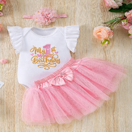 Baby outfit with 'My 1st Birthday' shirt and pink tutu skirt on a wooden surface with flowers.