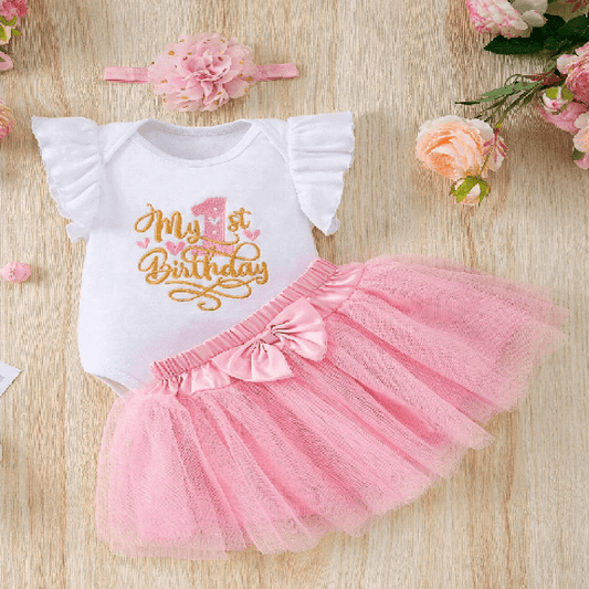 Baby outfit with 'My 1st Birthday' shirt and pink tutu skirt on a wooden surface with flowers.