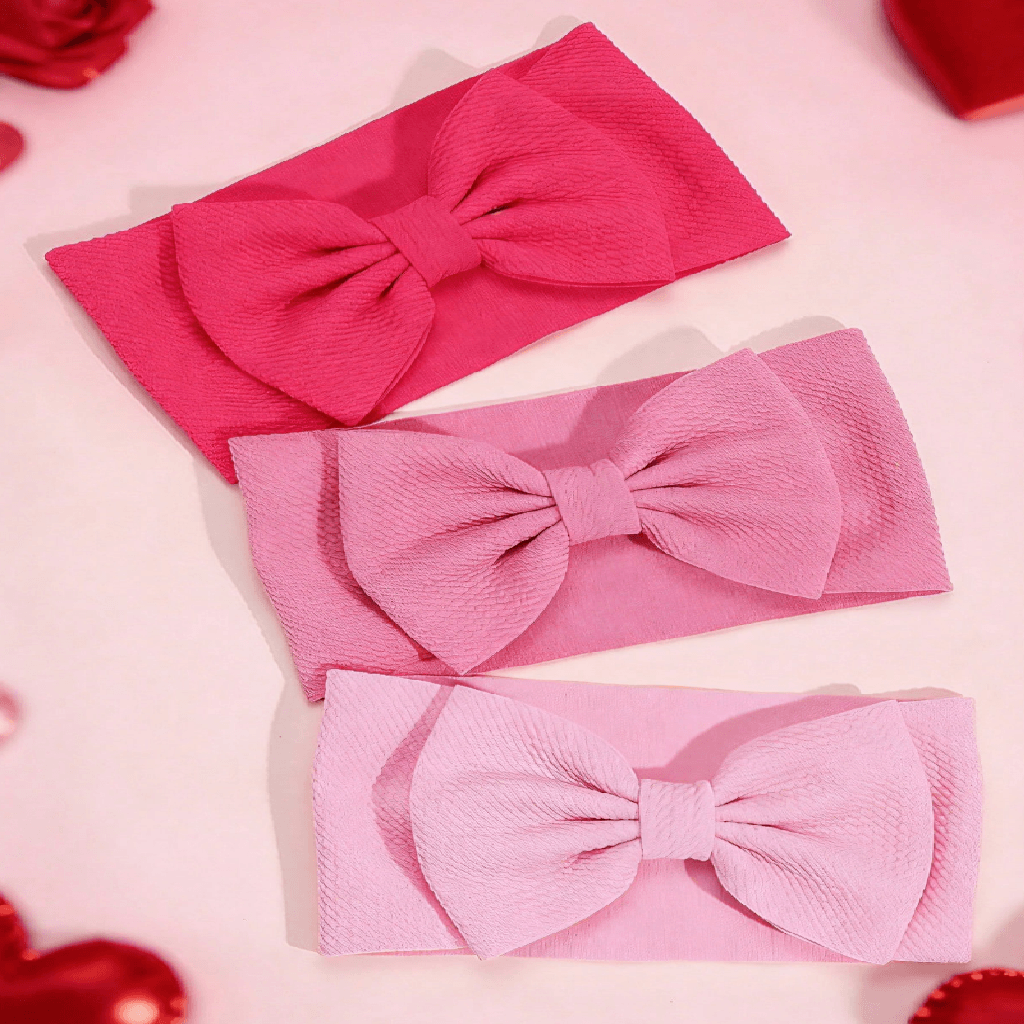 Three fabric bow headbands in pink shades, arranged diagonally on a light pink background with red decorative elements in the corners.