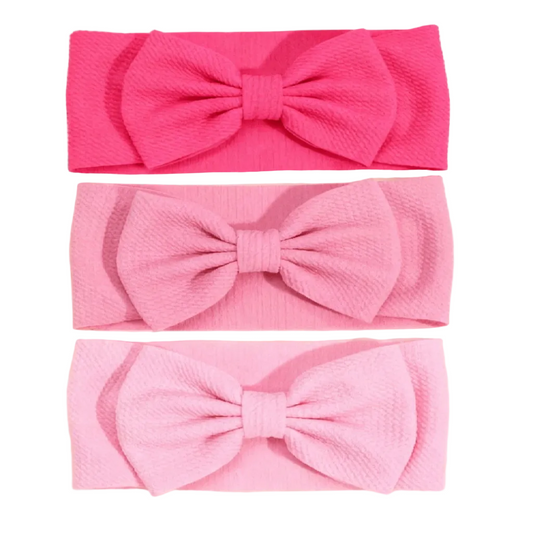 Three textured fabric headbands with large bows, shown in hot pink, light pink, and pale pink, arranged vertically on a light cream background with decorative rose petals.