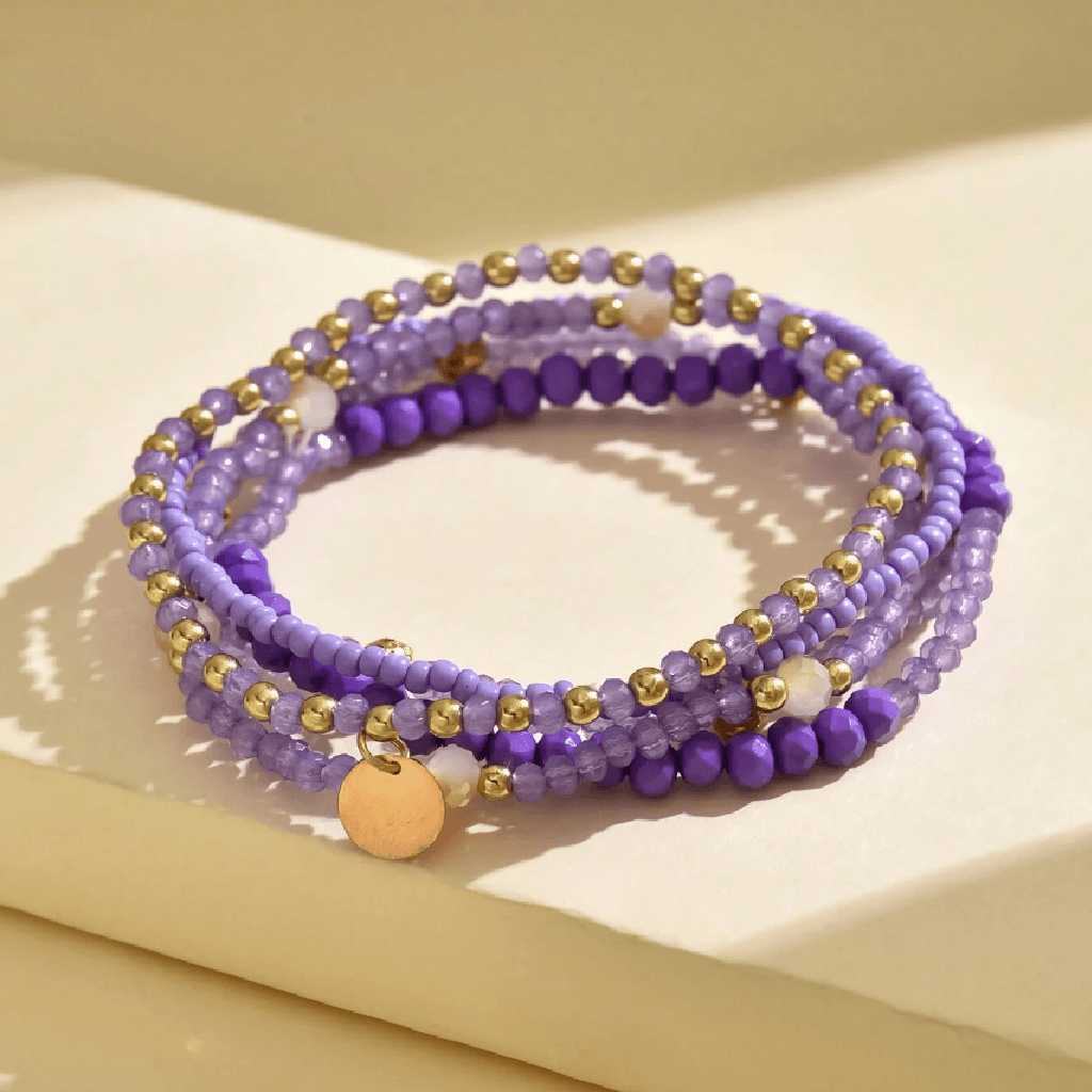 A set of three beaded stretch bracelets in shades of purple, accented with small gold beads and a round gold disc charm, displayed on a beige surface.