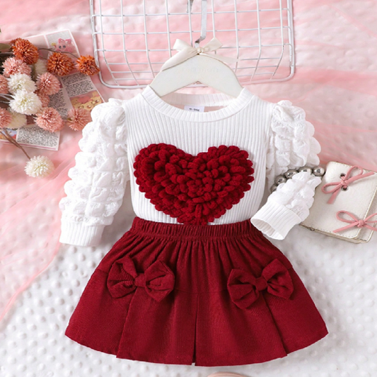 Children's outfit with white top and red skirt featuring a heart design on a pink background.