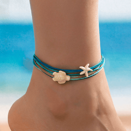 Anchored bracelet with turtle and starfish charms on a person's ankle against a beach background