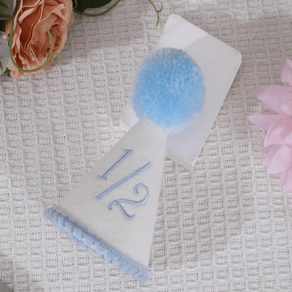 A white party table number card with a light blue pom-pom and a light blue embroidered '1/2' on a textured white fabric background with decorative flowers.
