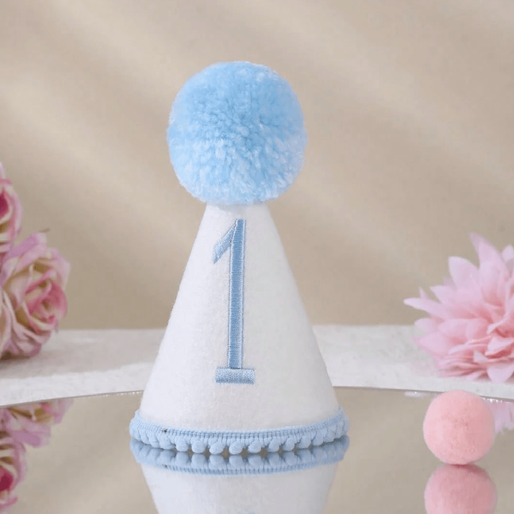 A white party hat with a light blue pom-pom, a light blue embroidered number 1 on the front, and a light blue pom-pom trim at the base, displayed on a reflective surface with pink flowers in the background.