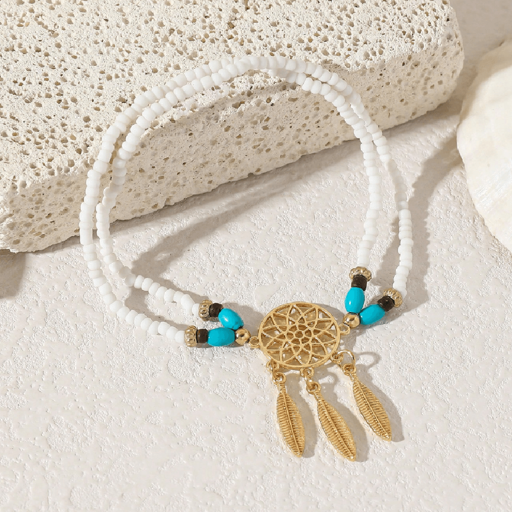 A bohemian-style double-strand beaded bracelet with a gold dreamcatcher charm, turquoise beads, and feather pendants, styled on a textured white surface with a decorative stone prop.