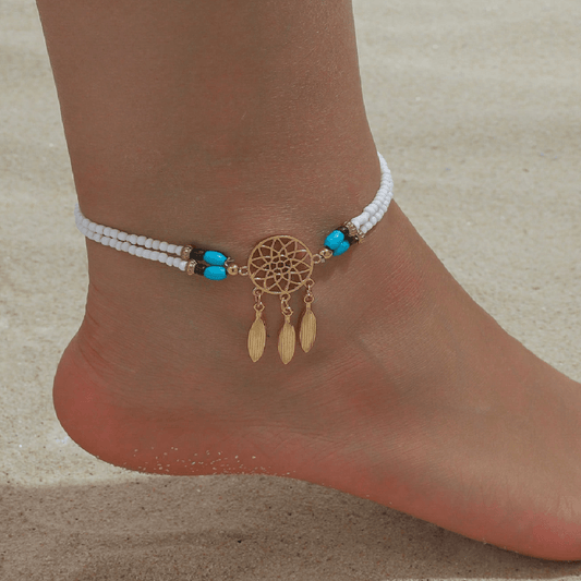 A close-up of a person's ankle and foot wearing a bohemian-style anklet on a sandy beach. The anklet features white seed beads, small turquoise blue beads, and a gold dreamcatcher charm with three dangling gold feathers.