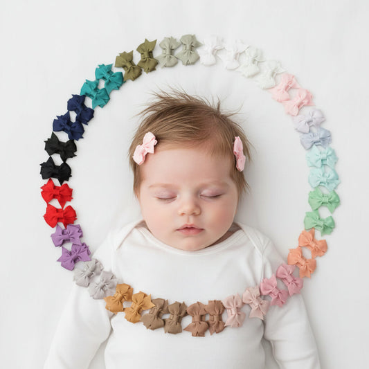 A sleeping baby wearing a white onesie lies on a white background, surrounded by a circular arrangement of small fabric bow clips in many colors. The baby has two matching pink bows in her hair.