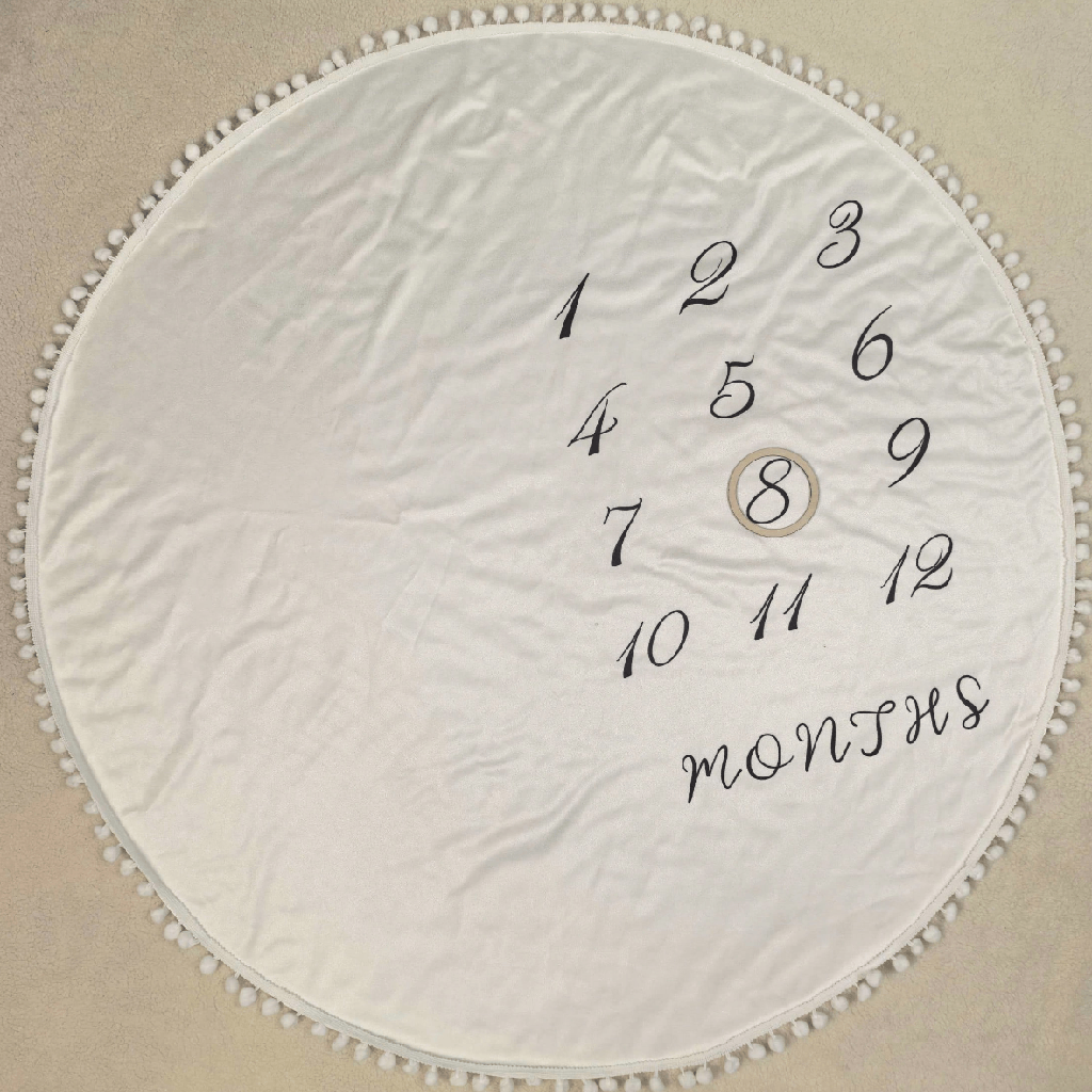 A round white fabric cover with a pom-pom trim, featuring printed black numbers 1 through 12 with the word 'months' and a circle containing the number 8 in the center, laid flat on a beige surface.